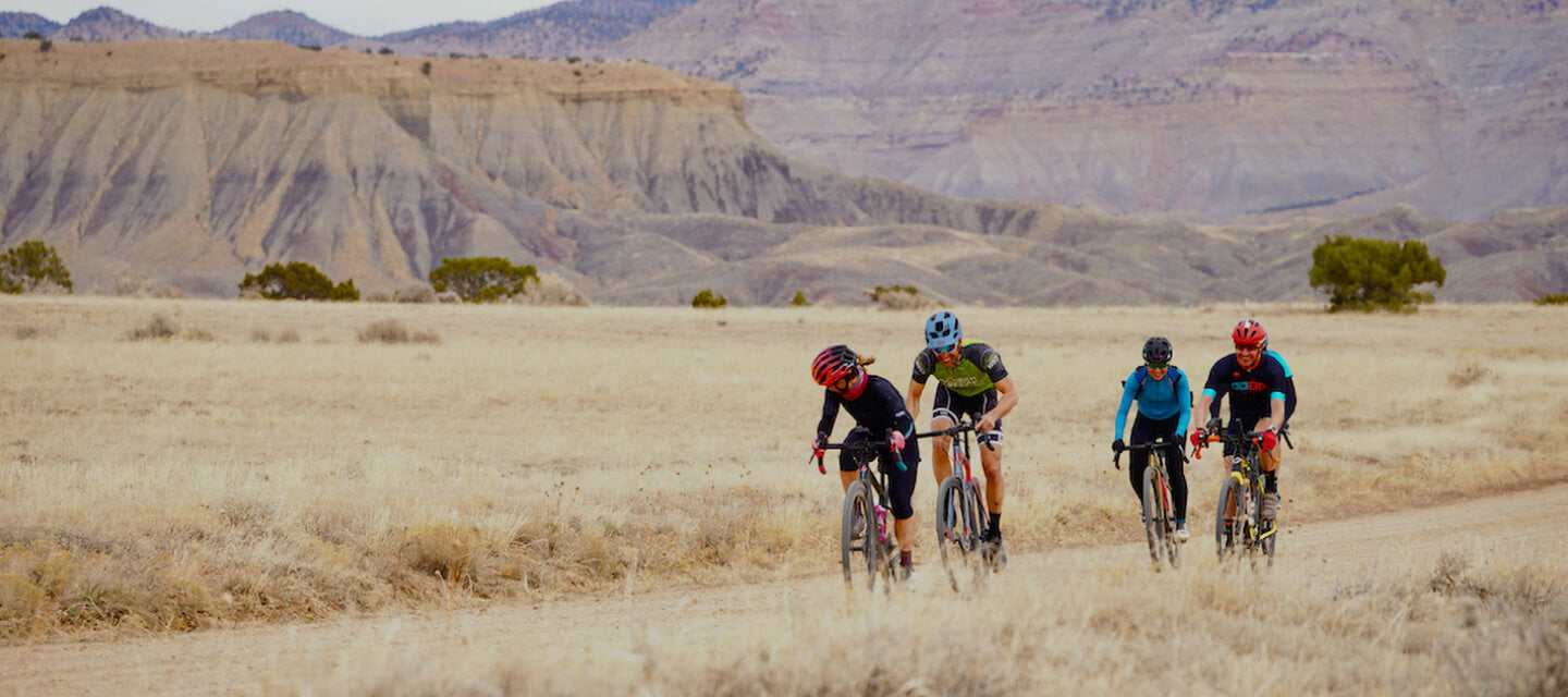 Group of cyclists riding gravel road in high elevation desert with hills and mountains in background