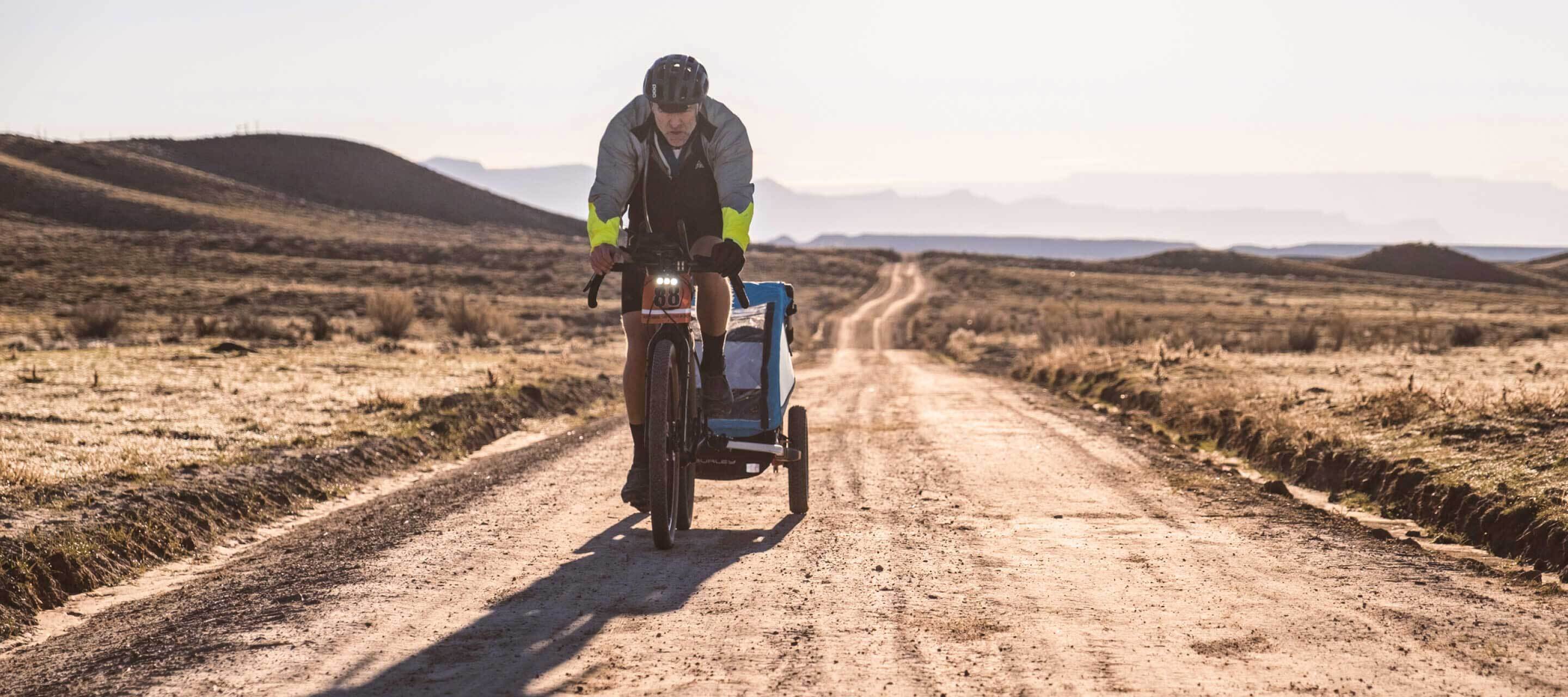 RJ riding bike on desert gravel road pulling kid trailer mountains in distance