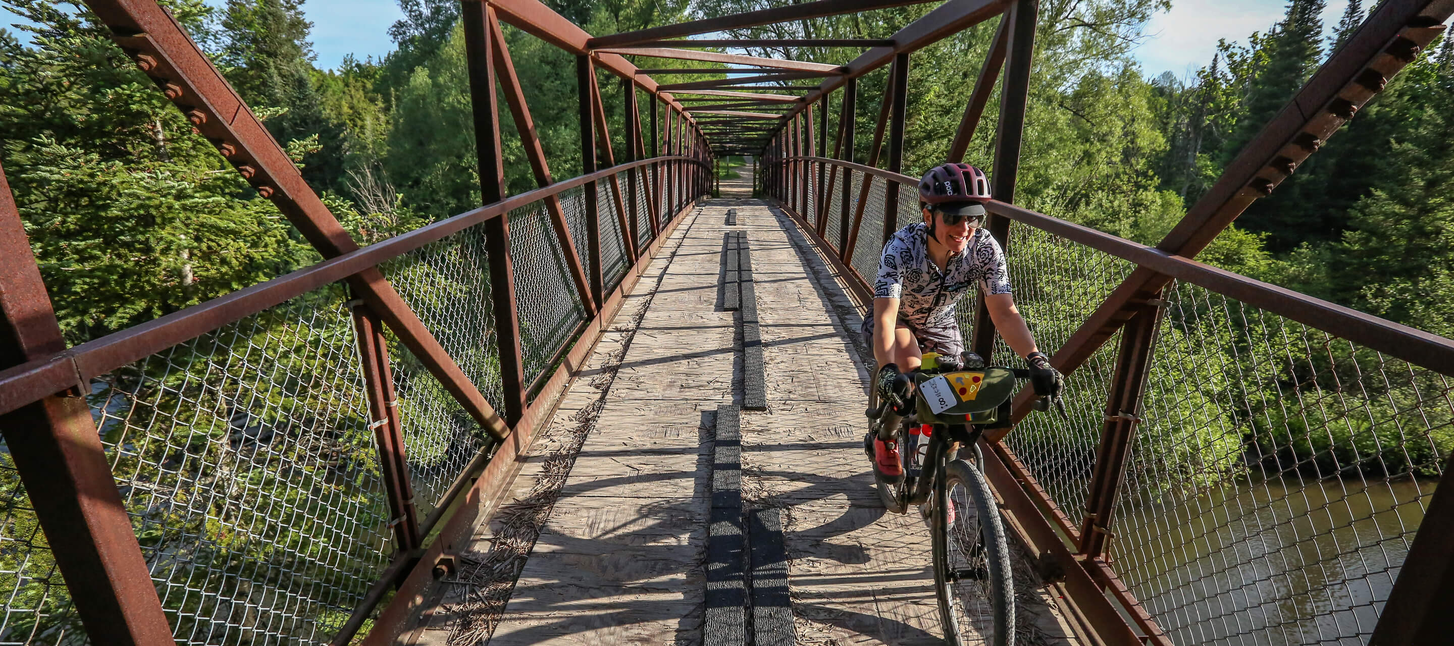 Jill smiling riding across narrow river bridge on drop bar bike with handlebar bag