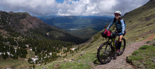 Dianna smiling while riding mountain bike loaded with gear on high elevation trail overlooking valley