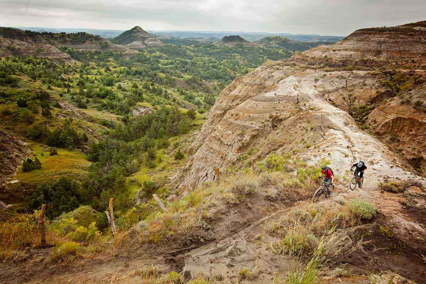 Two mountain bikers riding singletrack trail on hilly terrain in Badlands, mountains in background