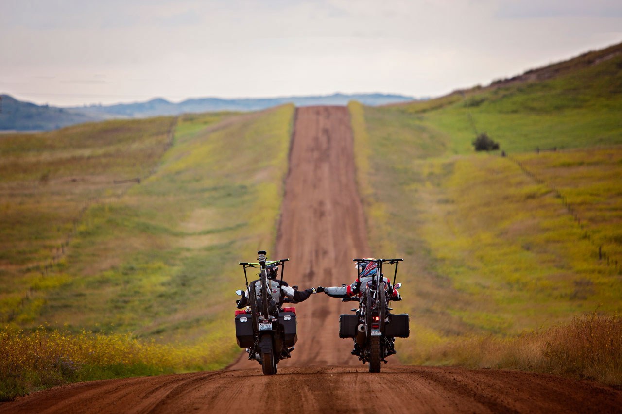 Two motorcyclists riding on gravel road with gear and mountain bikes loaded onto their motorcycles giving fist bump