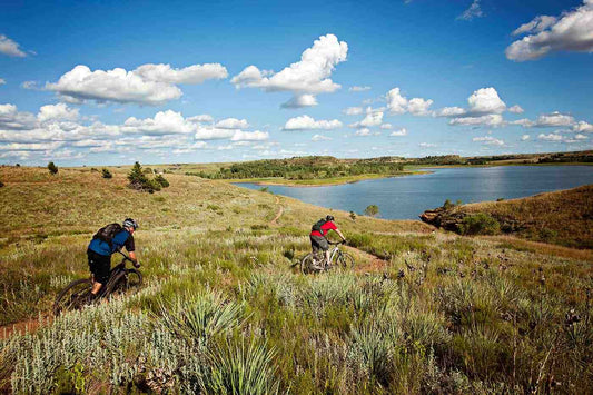 Two mountain bikers riding through open grassland trail on sunny day with lake, hills and trees in background