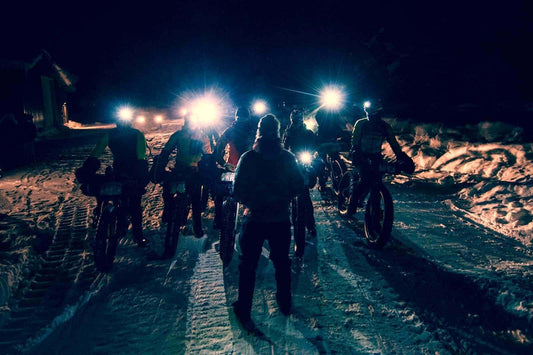 Rear view of a person facing a row of fat bike racing cyclists with headlamps on at night on a snowy road