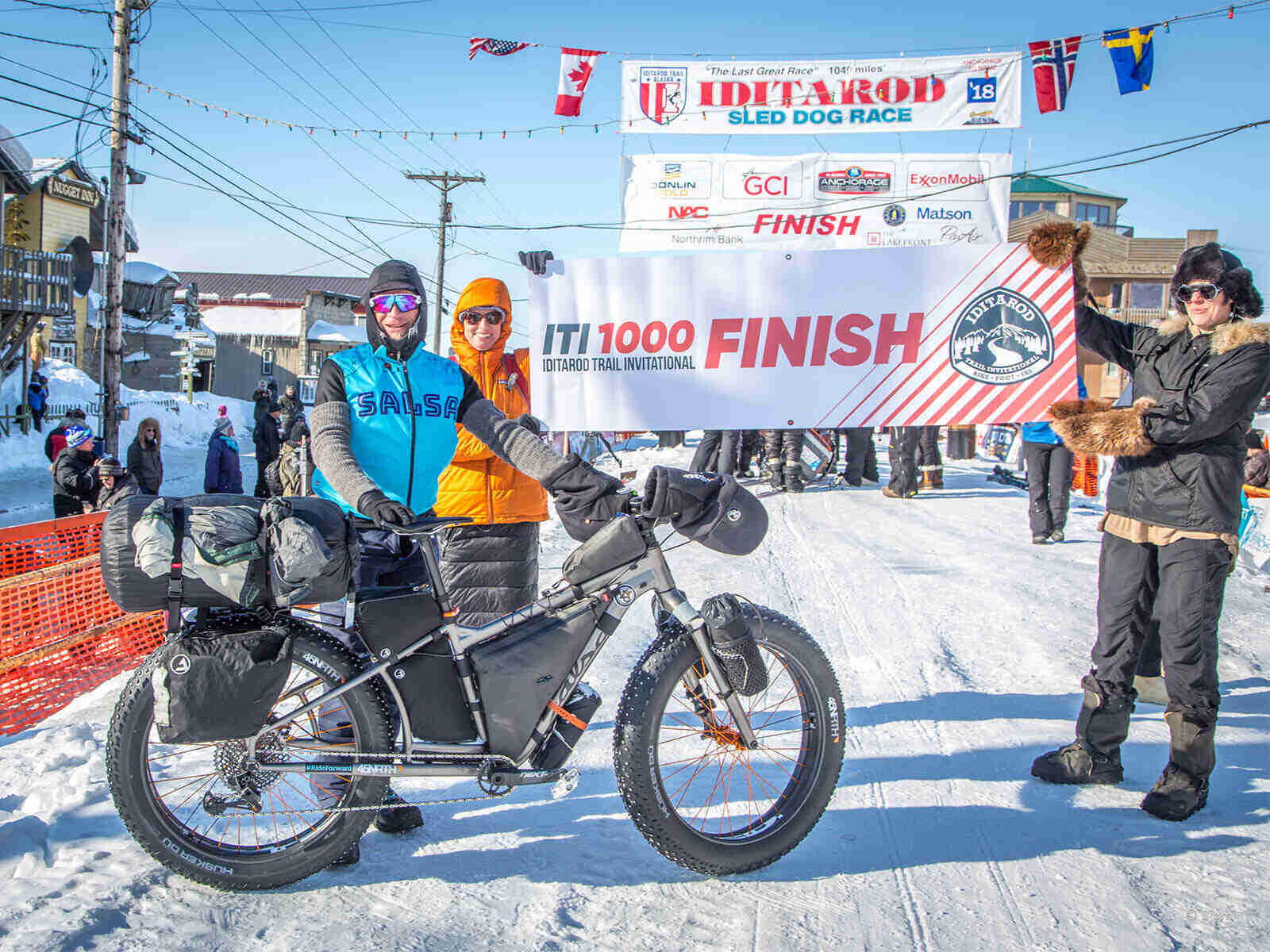 A winter cyclist posing with their fat bike, with 2 people holding up a "ITI 1000" sign, at a snow covered finish line