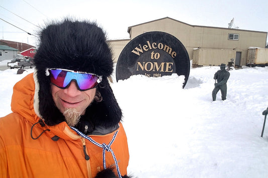 Front, chest up view of a person in winter attire, standing in a snow covered area with a "Welcome to Nome" sign behind