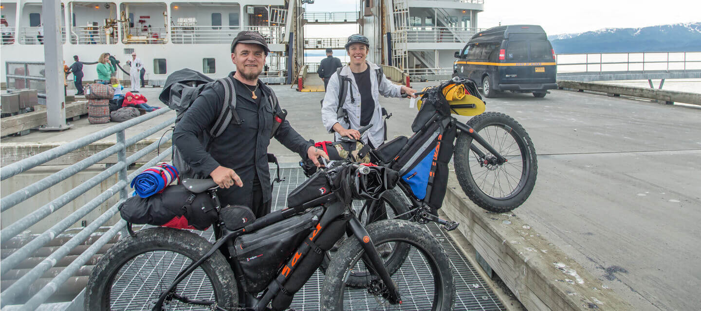 Kim and Bjorn standing behind their Salsa fat bikes on the left side, on a cement dock with a large ferry boat behind them