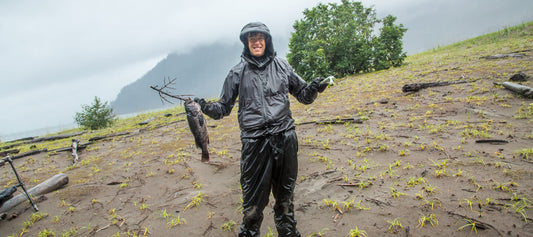 Kim dressed in rain gear, holding a rock fish in one hand, on a sandy shore with clouds above