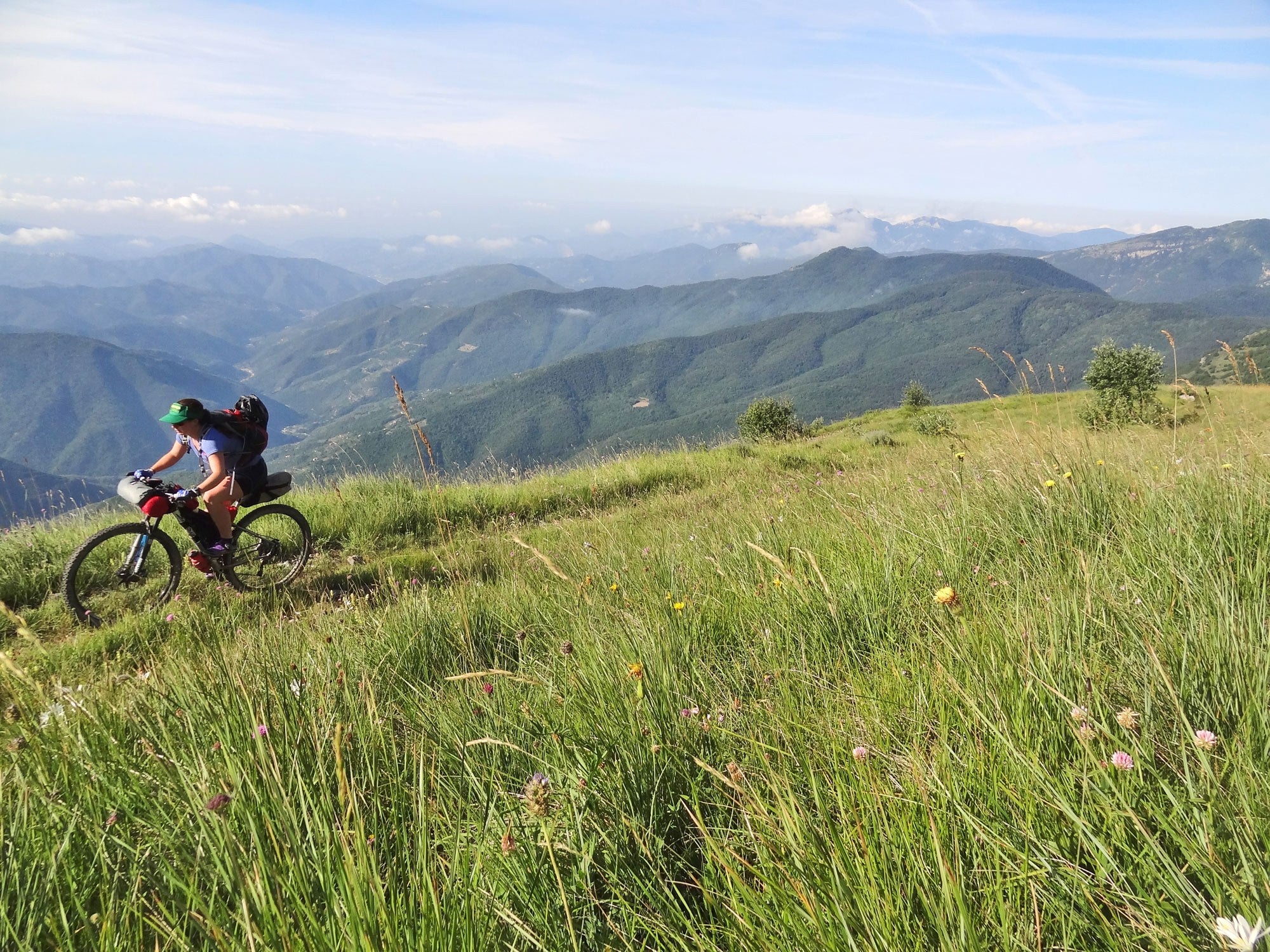 Kaitlyn climbing a grassy mountain alpine track on loaded mountain bike on sunny day with mountains in background