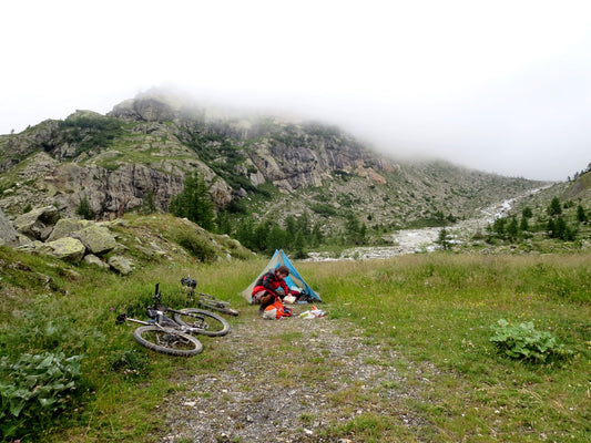 Kurt kneeling next to small tent packing backpack with two mountain bikes lying next to him, cloudy rocky ridge behind