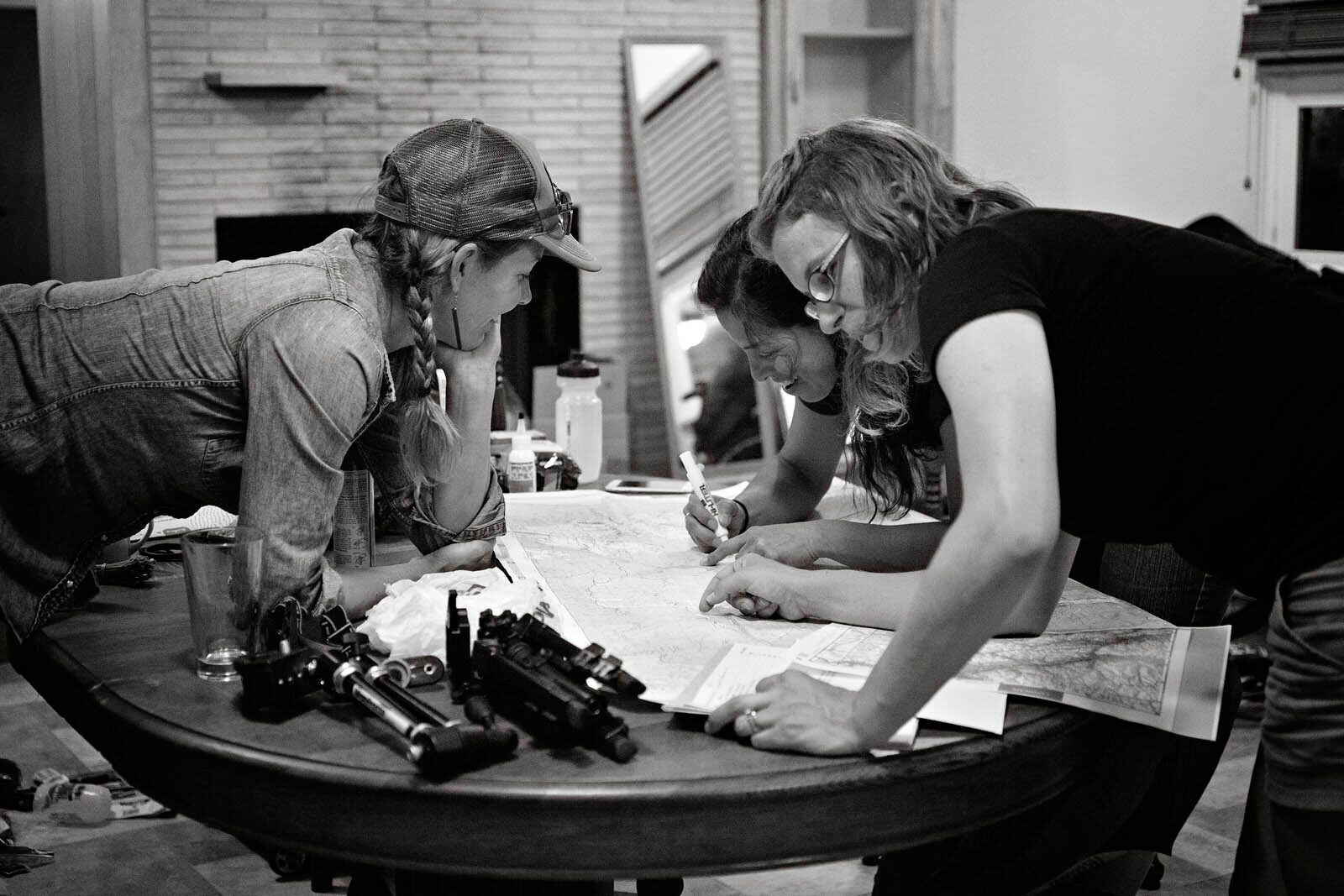 Kelly, Laura, and Trina inside home, standing leaning over table marking route on large paper map, gear scattered on table