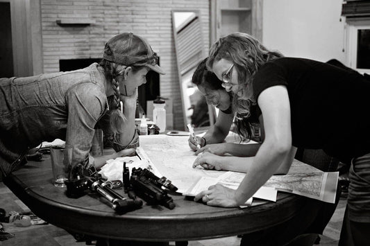 Kelly, Laura, and Trina inside home, standing leaning over table marking route on large paper map, gear scattered on table