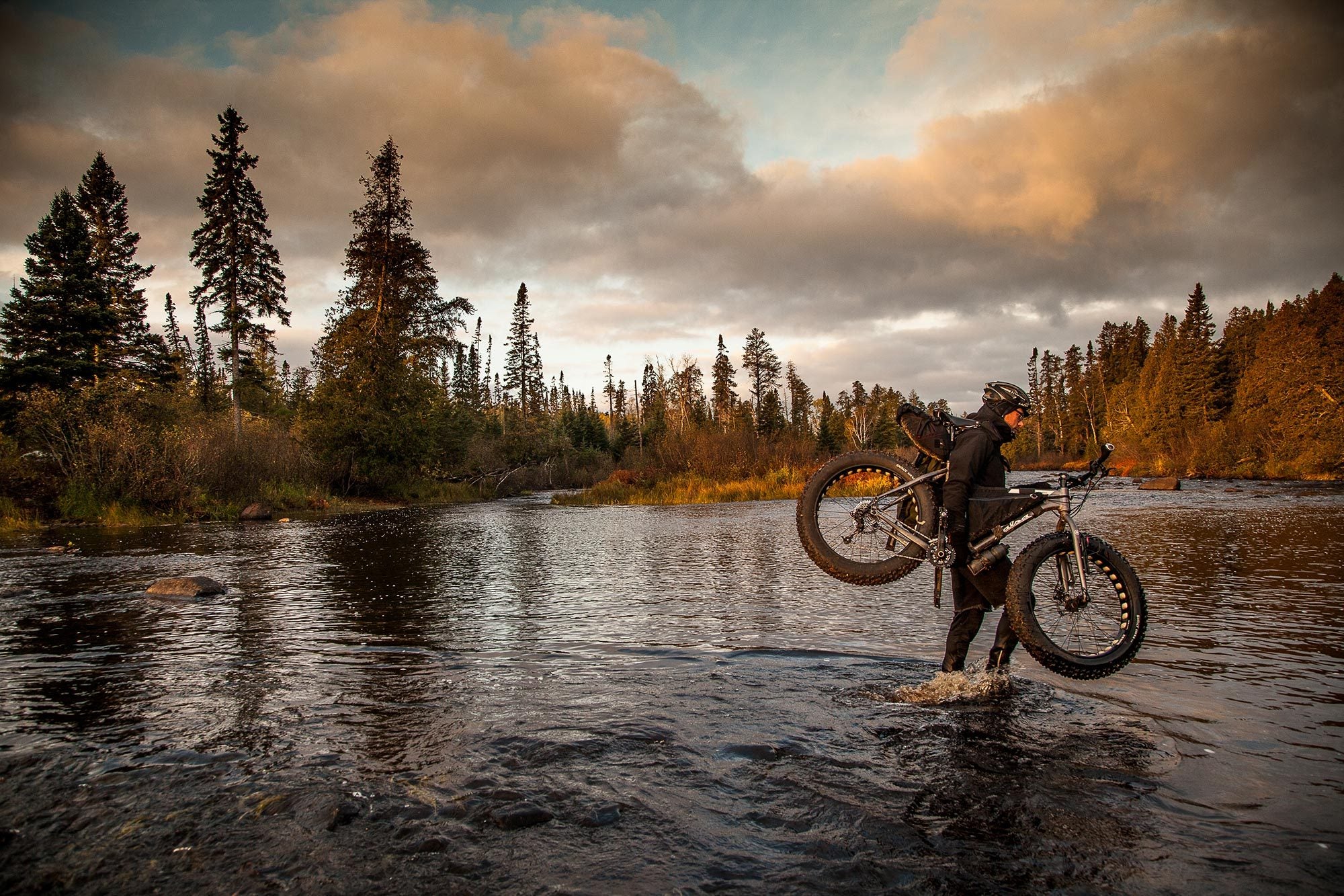 Mark ankle deep in the middle of a shallow river crossing wearing helmet, warm clothes and backpack, carrying fat bike