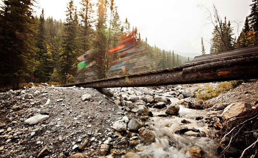 Two mountain bikers in a blur ride over bridge, rushing mountain stream filled with rocks below