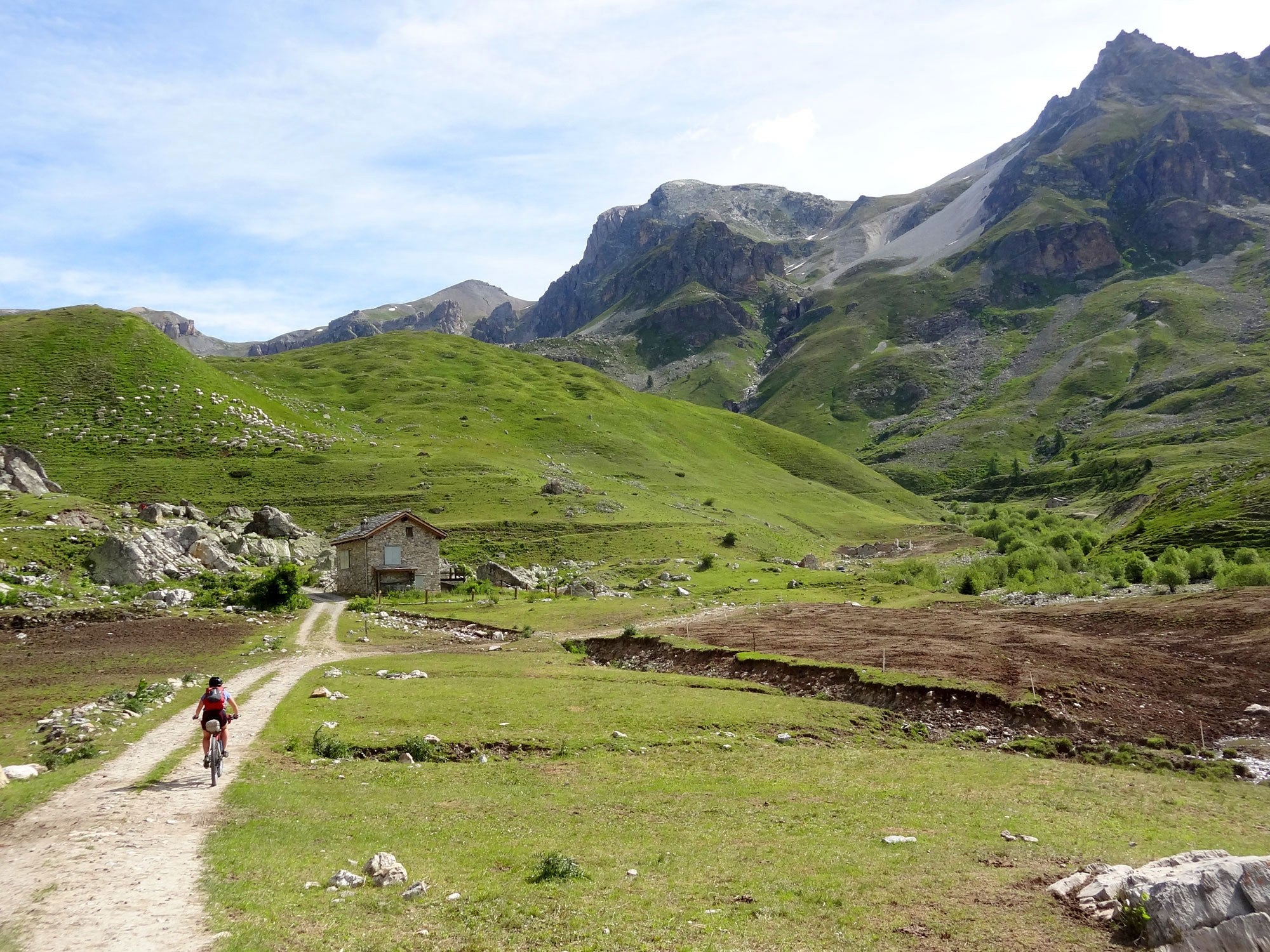 Kaitlyn riding loaded mountain bike on mountain double-track road leading to stone, grass hillside and mountains behind