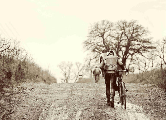 Cyclist pushing bike up muddy gravel road, cyclists riding out in front