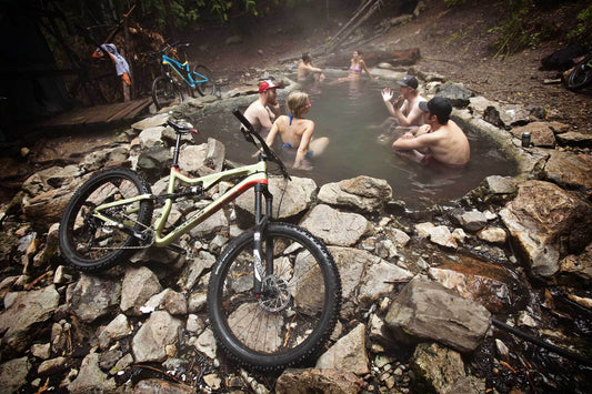 Bikes parked along outside of rock-lined hot spring pool in forest with people in swimsuits sitting in talking