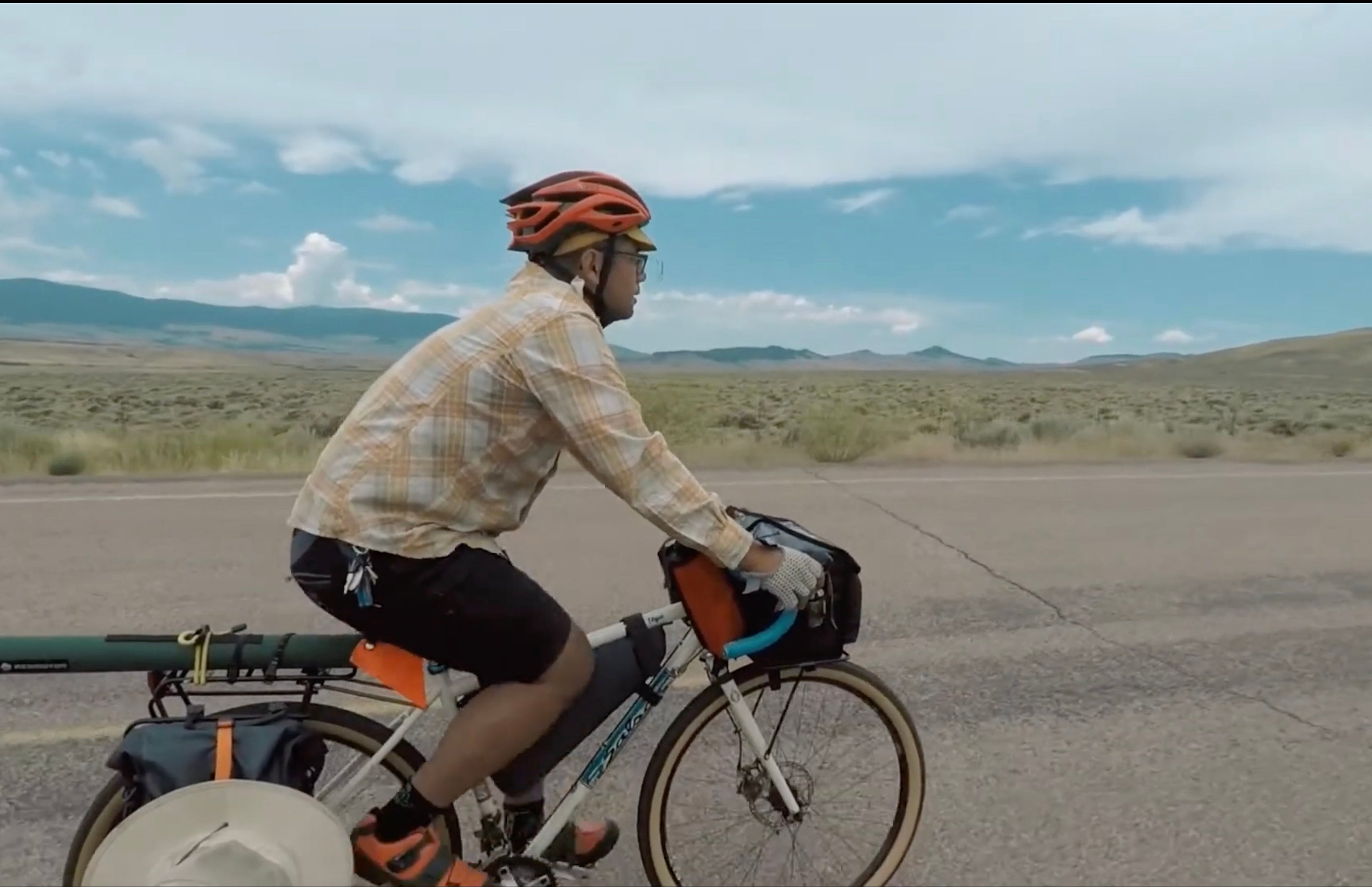 Cyclist rides a drop bar bike loaded with gear on a paved road along a desolate landscape.