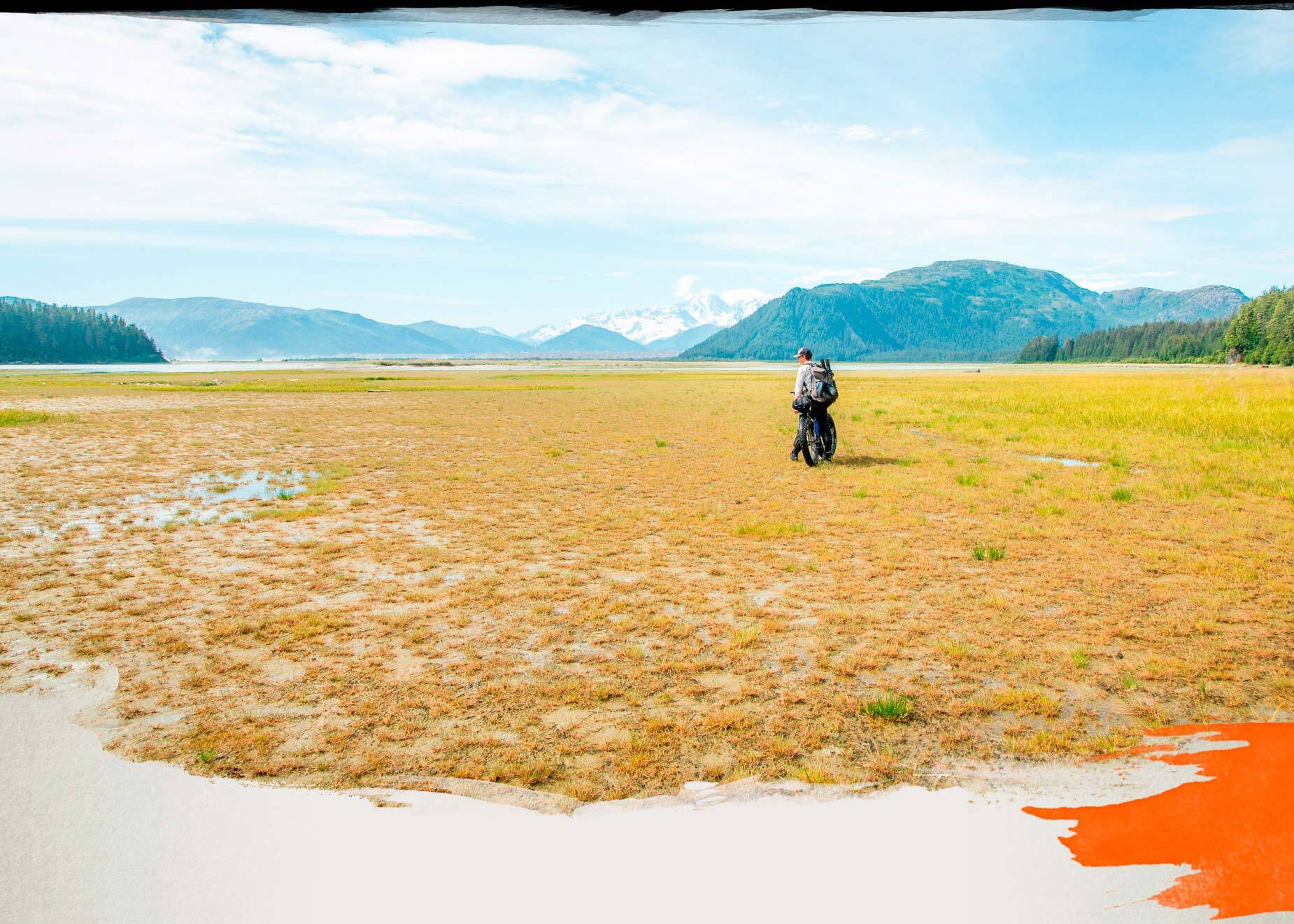 Kim stopped on loaded fat bike in marshy river valley, large mountains in distance