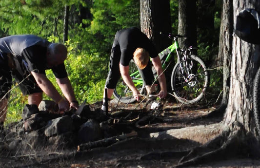 Cyclists bend over to gather their gear and load it back onto their bikes after camping in the woods.