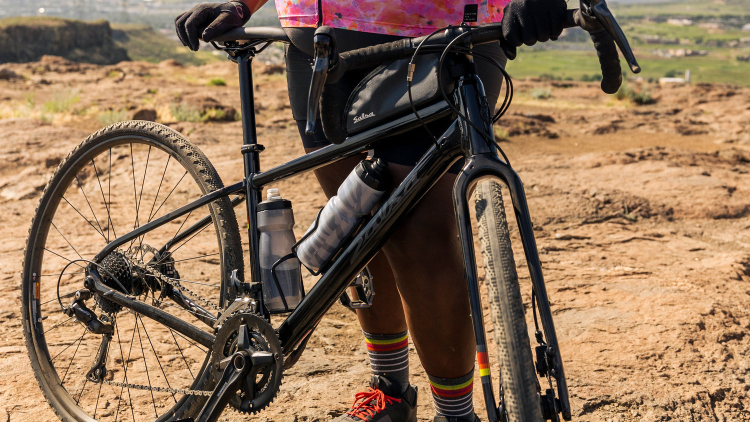 Cyclist stands on dry dirt terrain with a black Salsa Journeyer drop bar gravel bike.