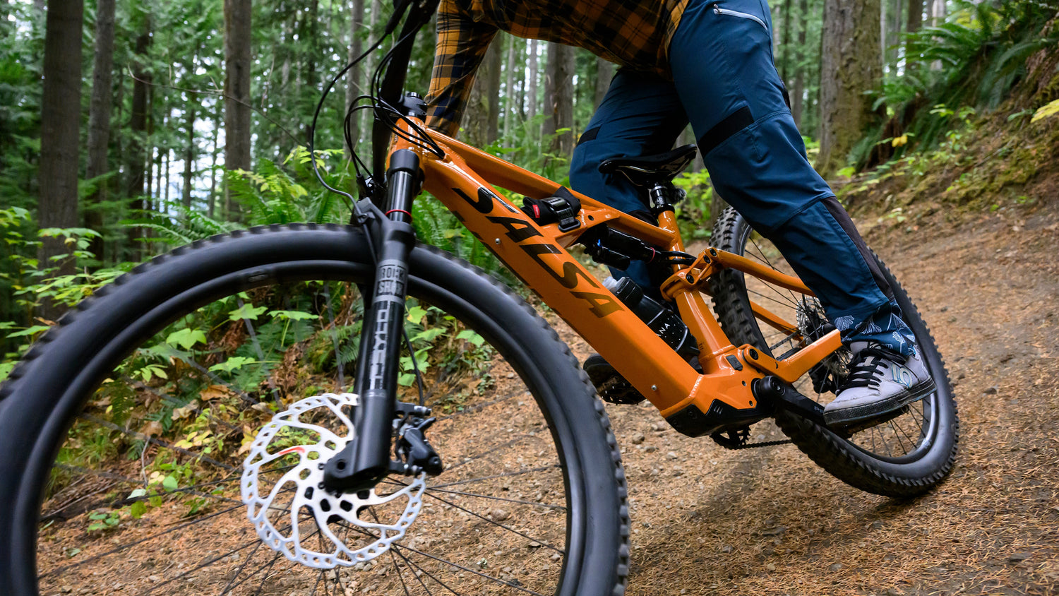 Cyclist rides an orange Salsa Moraine Ebike on dirt. Focus is on the fork and front wheel.