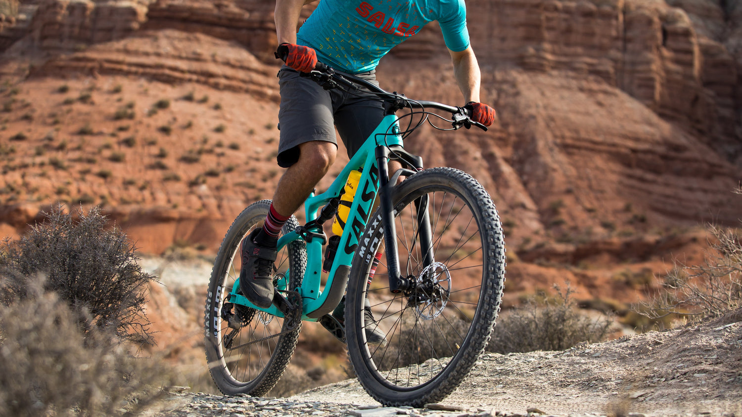 Mountain biker rides a turqoise Salsa Spearfish full suspension bike on a dirt trail.