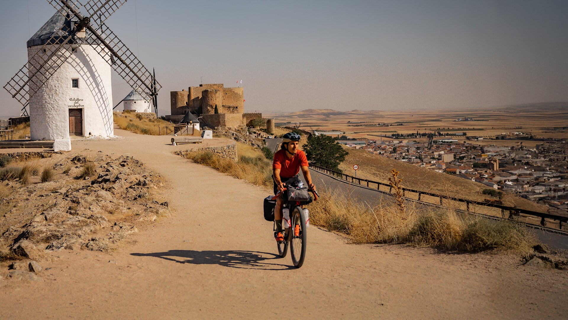 Cyclist rides a drop bar bike loaded with gear on a dusty road. A white windmill is behind them.