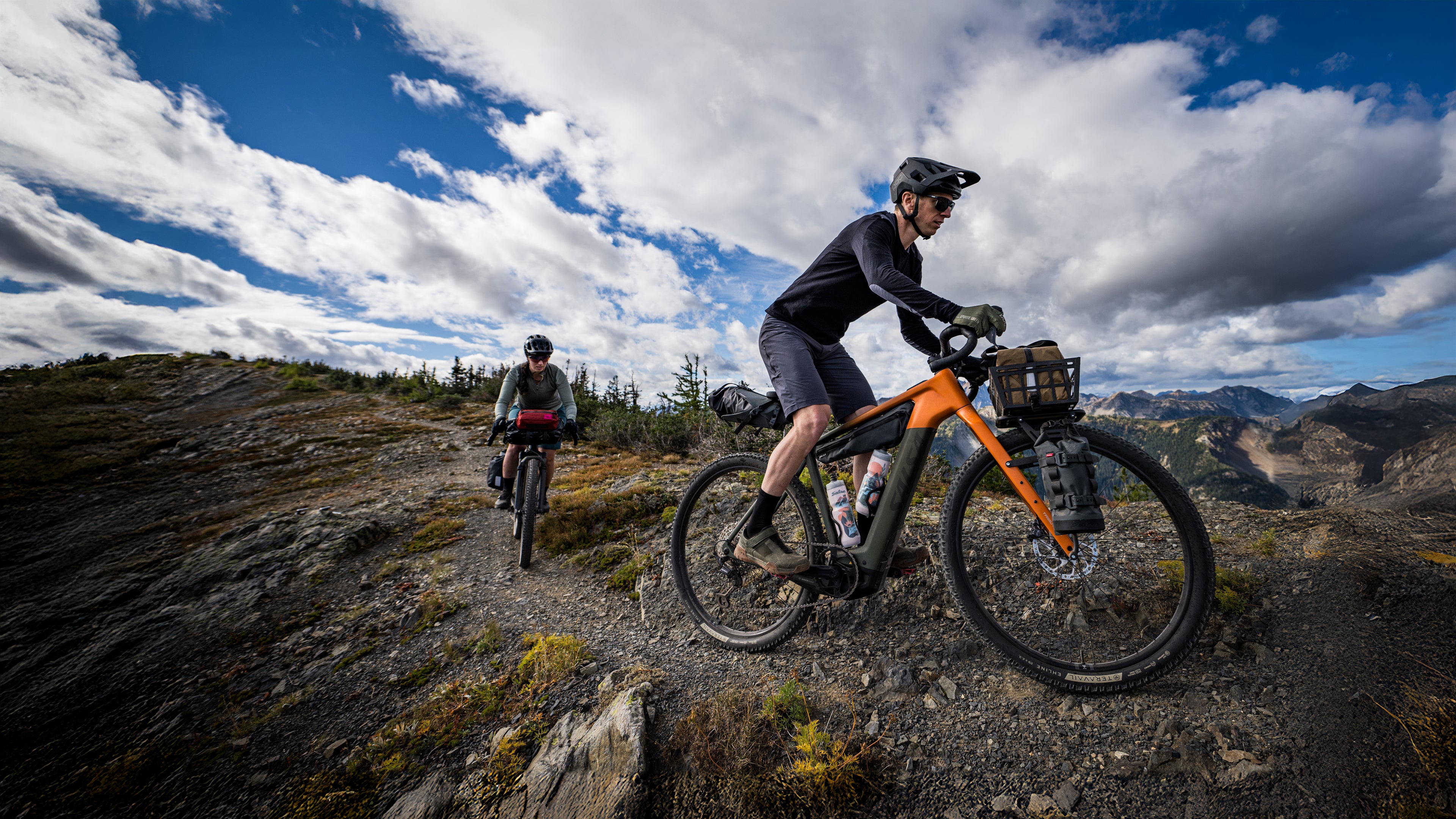 Two cyclists on Salsa Tributary ebikes ride on a narrow rocky trail in the mountains.