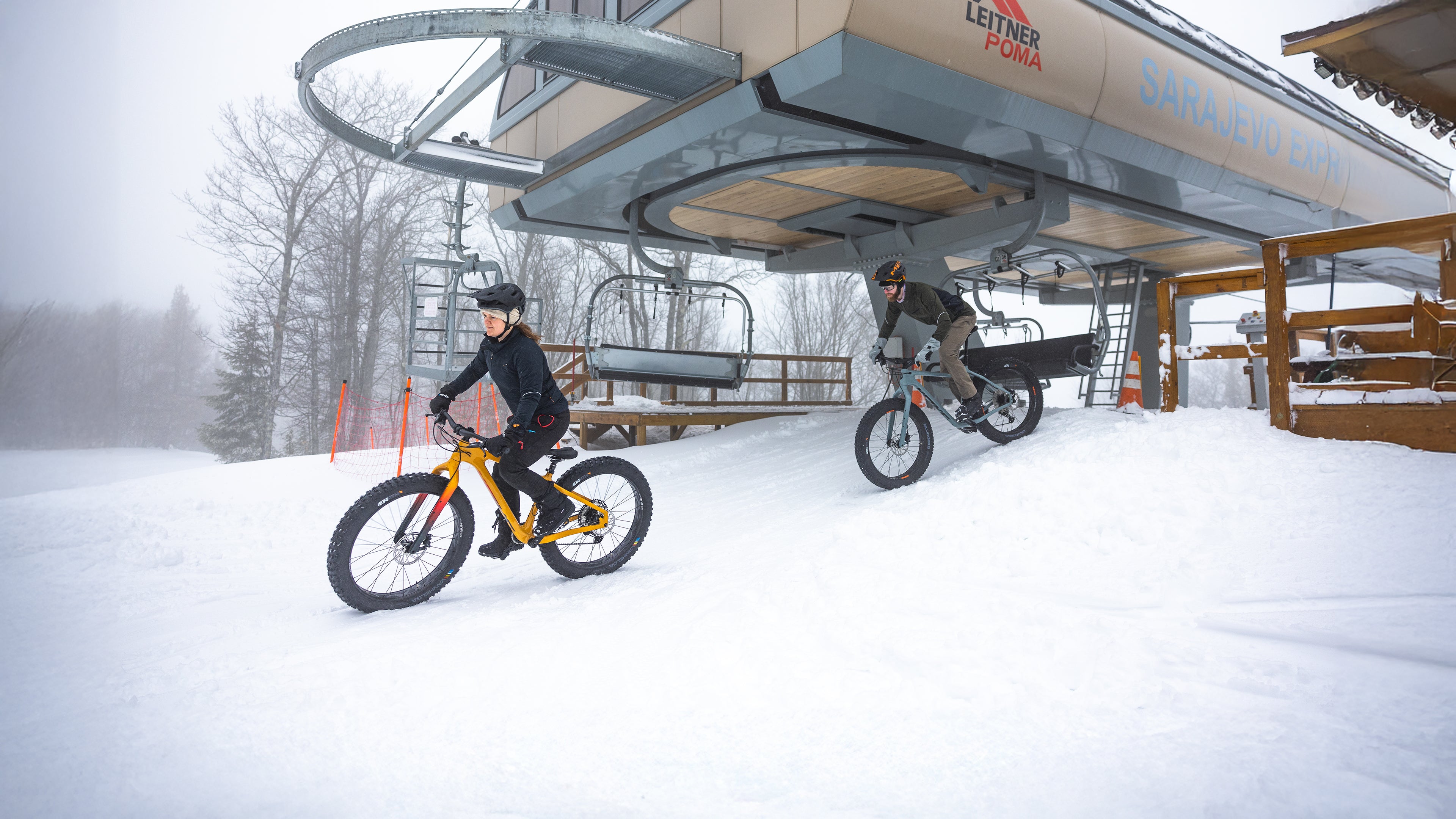 Two cyclists on fat tire bikes ride away from a chairlift in the snow.