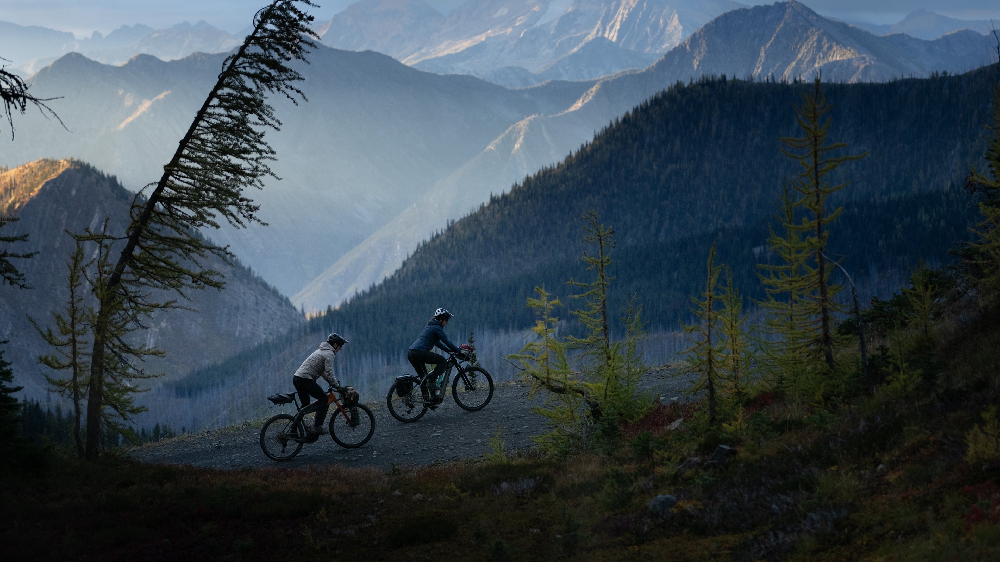 Two cyclists ride on an uphill slope over gravel. A mountain range sits behind them.